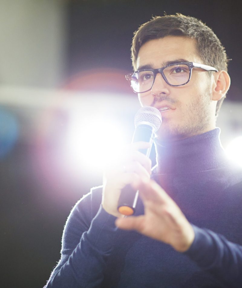 Head and shoulders portrait of confident young designer in eyeglasses holding microphone in hand while making presentation for his colleagues