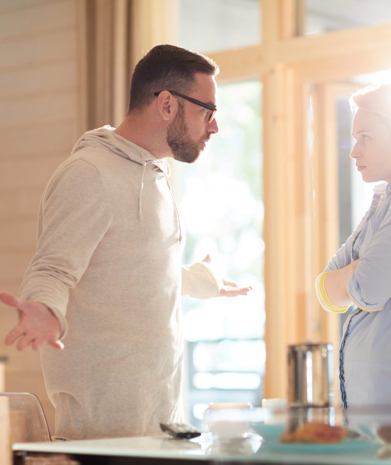 Young married Caucasian couple arguing about household duties while standing in home kitchen illuminated with bright morning sun